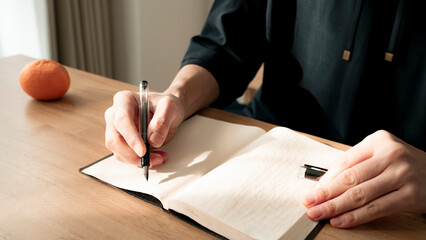 ノートに書き物をする男性の手元 思考の整理 ジャーナリング 丁寧な暮らし / Hands writing in notebook with pen and orange on table, journaling and planning concept