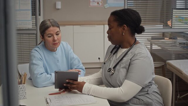 Female doctor and patient of different ethnicities reviewing digital medical test results together on tablet during scheduled clinic appointment