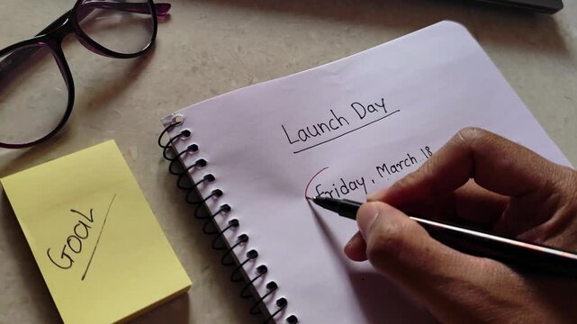 Close up video of a person writing and circling launch day date in a notebook with goal sticky note on desk. Business planning and startup product launch concept showing deadline scheduling.