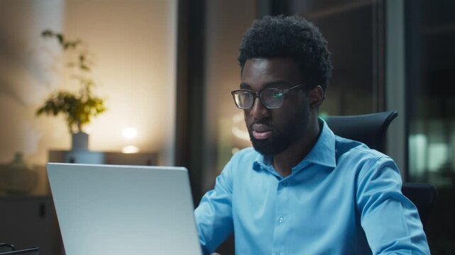 Focused African American male adjusting glasses while staring at laptop. Business shirt. Concentrating on complex task. Sitting upright at desk late night. Office interior glowing behind.
