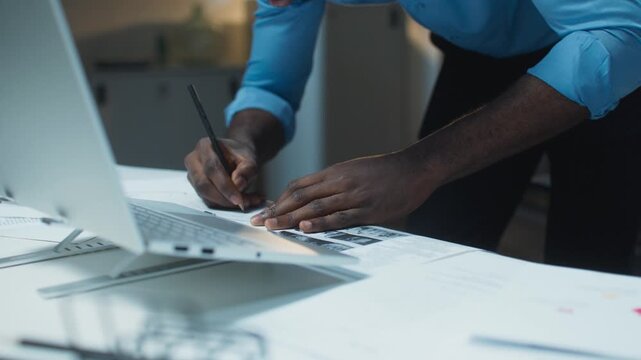 Close view of African American male hands writing notes. Business shirt sleeves rolled. Marking corrections on printed charts. Working beside open laptop late evening. Office desk scene.