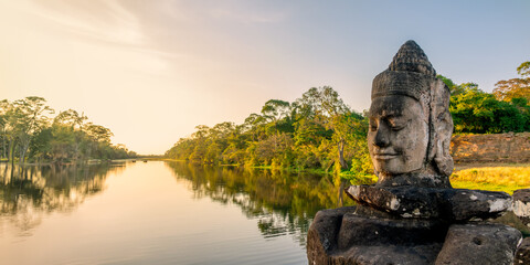 Obraz premium Deva head statue at Angkor Thom South gate, smiling serene Buddha face at sunset in Angkor, ancient moat with water reflections, Siem Reap, Cambodia panoramic header or web banner