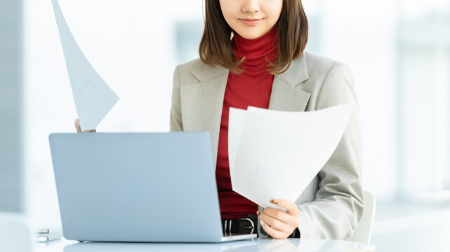 ノートパソコンを操作しながら資料を確認する女性ビジネスパーソン, Businesswoman reviewing documents while using laptop