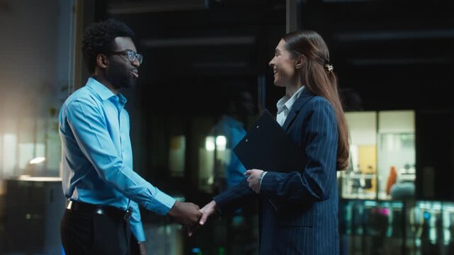 Business partners shaking hands after meeting near glass windows. Male smiling and thanking colleague. Female holding folder and agreeing on terms. Finishing cooperation discussion. Evening.