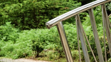 Fototapeta premium A close-up of a polished stainless steel garden railing against a lush green background of bushes and trees. This modern architectural detail provides both safety and aesthetic appeal.