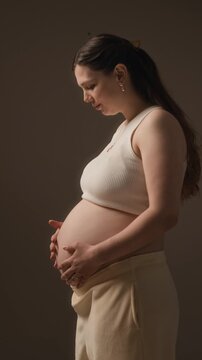 Vertical medium portrait of young Caucasian pregnant woman turning at camera, holding and caressing her bare belly in studio setting