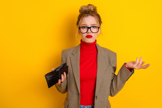 Young woman in stylish attire holding an open wallet in front of yellow background signifying confusion or financial concern
