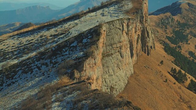 A striking natural landscape with a textured rock wall. A slope with patchy snow contrasts with a warm, golden hillside of dry grass. Majestic mountain scenery for travel or nature concepts