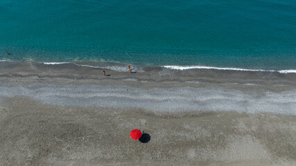 Obraz premium Minimalist aerial view of a wide pebble beach where a single red umbrella stands as a vibrant focal point. In the distance, near the shoreline, two small figures walk by the turquoise waves of the sea