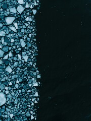 Aerial top-down view of fragmented sea ice floating along the edge of dark open water. Numerous irregular ice pieces of varying sizes cluster densely on one side, forming a textured mosaic. © Gala_Rocci