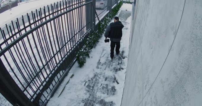 Security Camera Records Man Rushing Down Snow Cleared Sidewalk, Glancing Over Shoulder. Man Wears Dark Glasses, Black Cap, and Carries Leather Bag Tense Atmosphere and Security Concern. Back View.