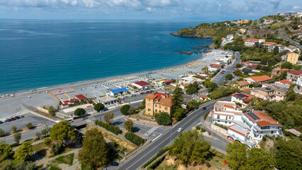 Aerial panorama of a Mediterranean coastline, featuring a vast pebble beach with organized lidos and parking lots, leading toward a rocky promontory. It is the beachfront of Scalea, in Calabria, Italy