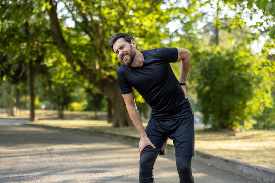 A young male athlete stands in the park, bent over due to back pain, holding his lower back with his hand