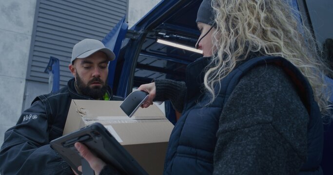 Logistics Team Unloads Parcels From Delivery Van During Snowfall. Man Moves Boxes While Woman Uses Scanner and Tablet Check Shipments. Coordinated Winter Delivery Operations in Cold Conditions.