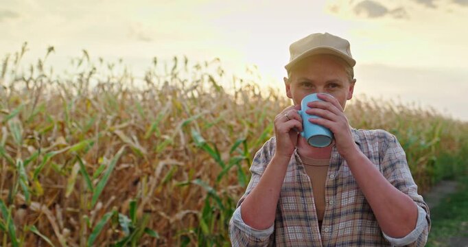 Middle-aged woman farmer standing in corn field at sunset, drinking hot tea from mug during work break, calm rural lifestyle and agriculture concept