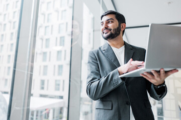 Fototapeta premium Man holds laptop while standing by a large window in a modern office space during daytime