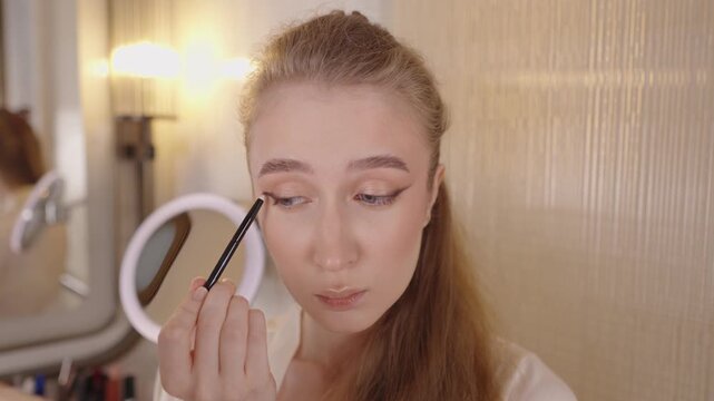 Beautiful young woman concentrating while applying classic winged eyeliner with a black pencil, looking in a round mirror as part of her daily beauty routine in a well lit bathroom