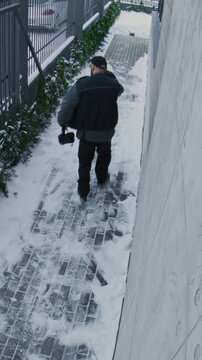 Security Camera Records Man Rushing Down Snow Cleared Sidewalk, Glancing Over Shoulder. Man Wears Dark Glasses, Black Cap, and Carries Leather Bag Tense Atmosphere and Security Concern. Vertical Shot.