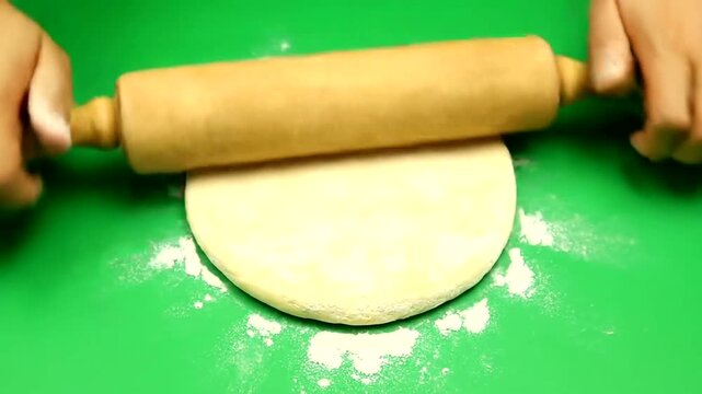 Hands rolling out dough with a wooden rolling pin on a green surface.
