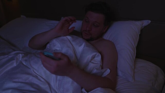 Sleepy man lying in bed at night using his phone, feeling shocked and upset after reading bad news online, suffering from anxiety, depression, and doomscrolling addiction