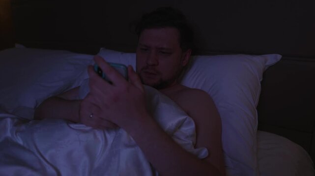 Concentrated young man scrolling and swiping on his smartphone while lying in bed late at night, suffering from insomnia, screen addiction in a dark bedroom before sleeping