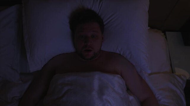 Overhead view of frightened man suddenly jolting awake in the middle of the night, breathing heavily and scanning the dark bedroom after disturbing dream, gasping for air and looking around