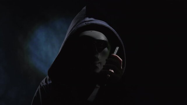 Telephone scammer wearing black mask, hiding face, using smartphone to commit fraud, speaks on mobile phone against dark background with backlighting