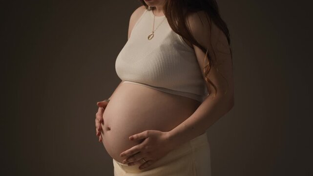 Cropped portrait of Caucasian pregnant woman gently holding her baby bump during maternity photoshoot, highlighting pregnancy, motherhood and prenatal wellness