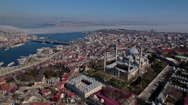 Aerial drone view of S&uuml;leymaniye Mosque in Istanbul, featuring its domes and minarets with the Golden Horn, bridges, boats and historic city skyline in the background.