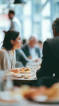 A professional woman and an older man share a meal at a stylish restaurant, discussing work matters. The atmosphere is bright, with a blurred background of other diners and large windows