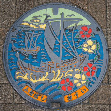 Tokyo, Japan - Oct 16 2024, close-up view of a manhole cover decorated with a image of two ships against the sea and flowers of a camellia tree, near the Funabashi metro station, Tokyo, Japan