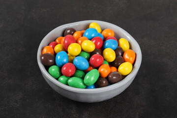 Dragees with peanuts and milk chocolate, covered with a crispy multicolored glaze. In a bowl. Dark gray background. Close-up