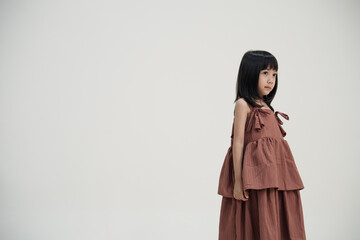 Asian girl in brown dress standing sideways on neutral background, cute children fashion portrait.