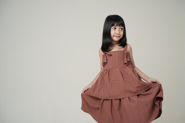 Naklejka premium Smiling Asian girl in brown dress holding skirt on neutral background, cheerful child fashion pose.