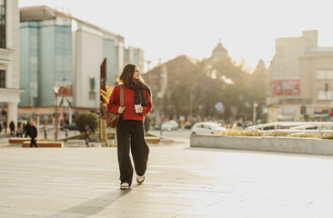 Smiling young woman walking down the sunny European street with cup of coffee to go