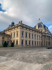 Obraz premium Majestic historic building with classical columns and a domed roof under a cloudy blue sky in Paris. Wide stone square and elegant French architecture featuring the national flag.