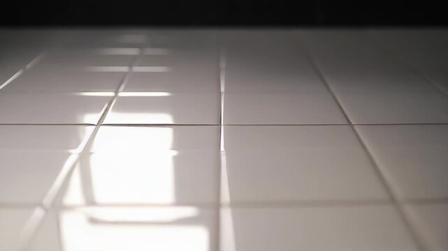 Bright light illuminates a clean, minimalist tiled floor, creating geometric patterns and reflections across the smooth, shiny surface, highlighting a sense of cleanliness