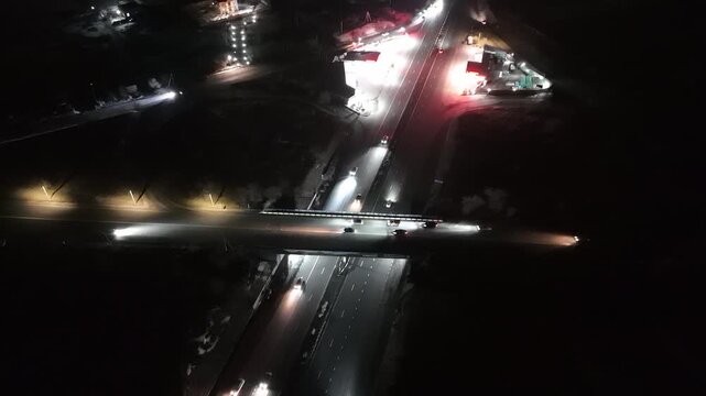 Aerial view of a highway interchange at night with car traffic, featuring cinematic film grain texture and atmospheric lighting in deep darkness.