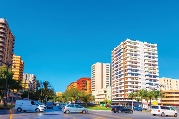 Street scene in Malaga features prominent residential architecture. Vehicles navigate busy urban intersection surrounded by colorful high rise buildings and palms