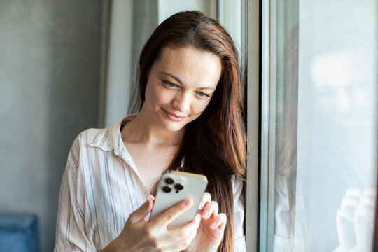 Smiling woman using smartphone by window at home