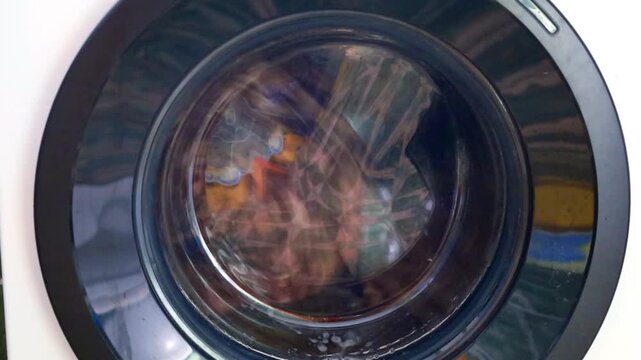 Rhythmic rotation of a washing machine drum during the laundry cycle. Colorful clothes mix in soapy water through the glass door. Modern household appliance gently cleans textiles,