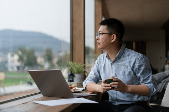 A man wearing glasses works on a laptop by the window with mountain view