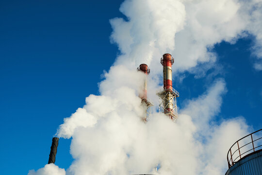 Two chimneys of a natural gas heating plant partially hidden by dense white steam. Water vapor and condensation cloud against a blue sky, winter utility background.