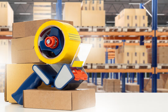 Warehouse, logistics center. Packaging tape dispenser seals cardboard boxes on packing table, parcel preparation for shipping in storage facility.
