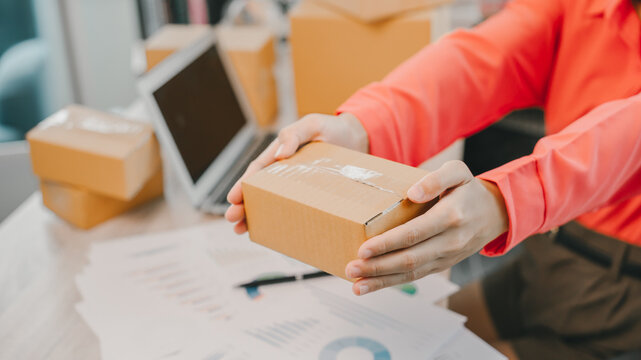 Online business owner. Small cardboard box hands holding box office desk laptop computer business documents shipping package person orange shirt delivery parcel