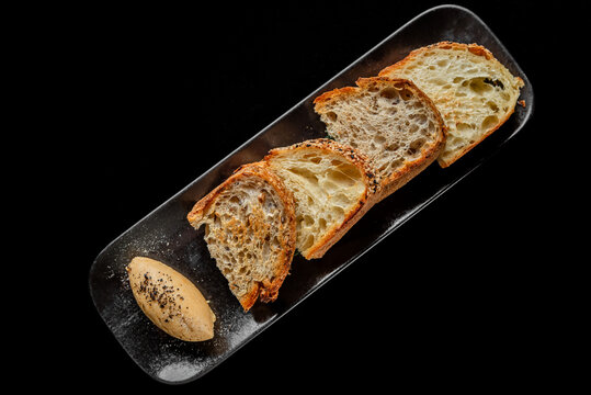Slices of crusty artisan sourdough bread and rye bread arranged on a dark plate with a quenelle of whipped butter, shot from a top-down perspective against a black background.