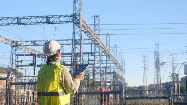 Female engineer in safety vest and hard hat using a digital tablet, monitoring an electrical substation for energy grid management and infrastructure maintenance