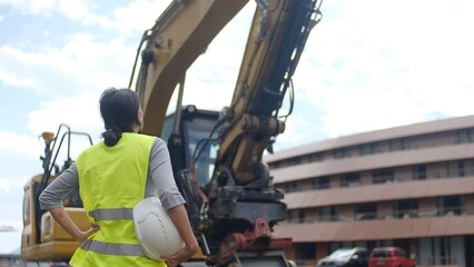 Female project manager overseeing a construction site, holding a hard hat, wearing a safety vest, and looking up at an excavator against an urban background © DawDunia