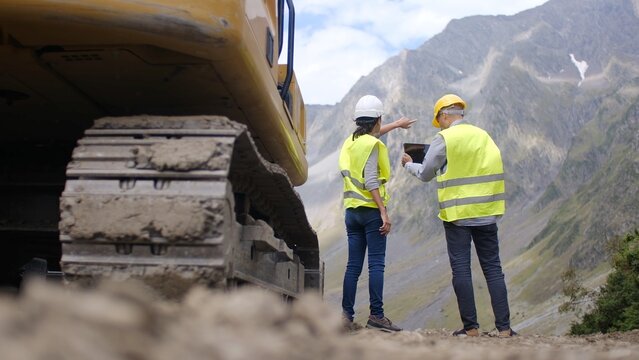 Construction workers and engineers discussing project plans using a digital tablet at a mountain road building site, overseeing heavy machinery and earthmoving operations