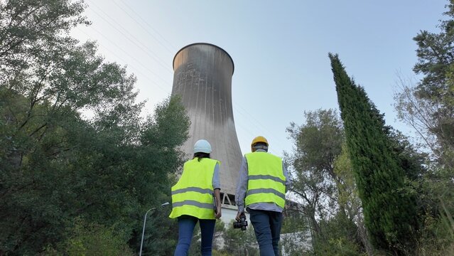 Engineers and technicians in hard hats and reflective vests walk toward a large cooling tower at a power plant, inspecting infrastructure and monitoring operational efficiency for sustainability